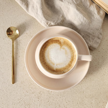 Vedere de sus a unei cești bej cu cafea, farfurioară și linguriță aurie